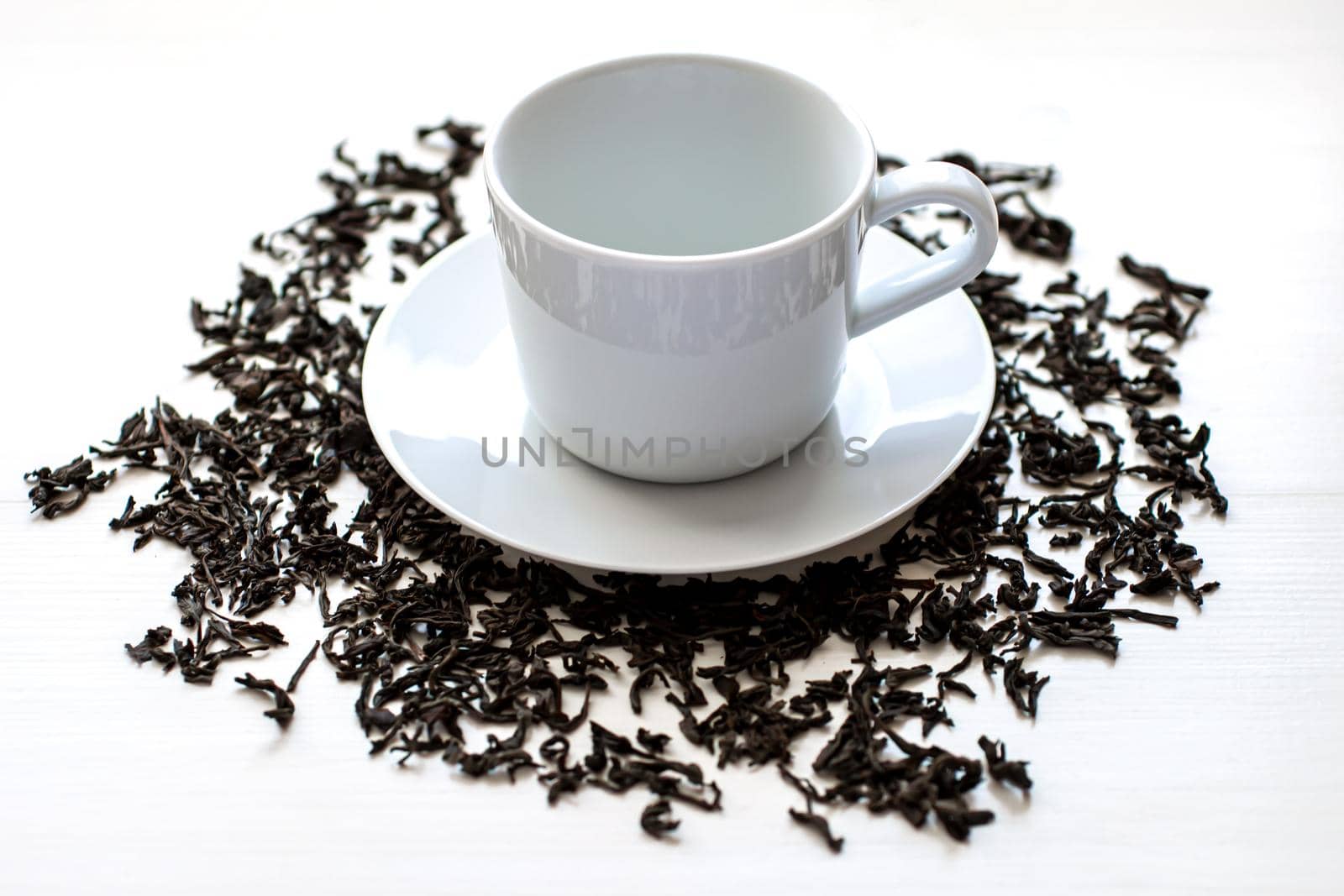 Ceramic white cup and saucer with scattered dried tea leaves around on a white wooden background