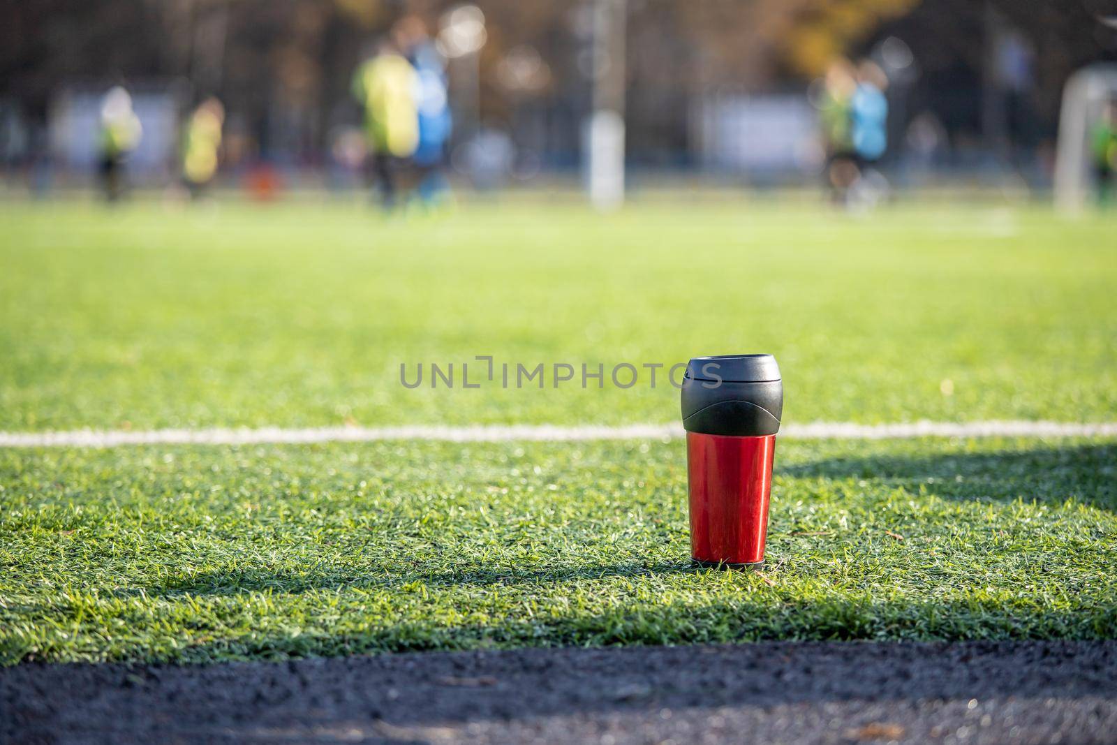 A red sports bottle with a black lid is located on a green football field made of synthetic grass. There are football players on a blurry field.