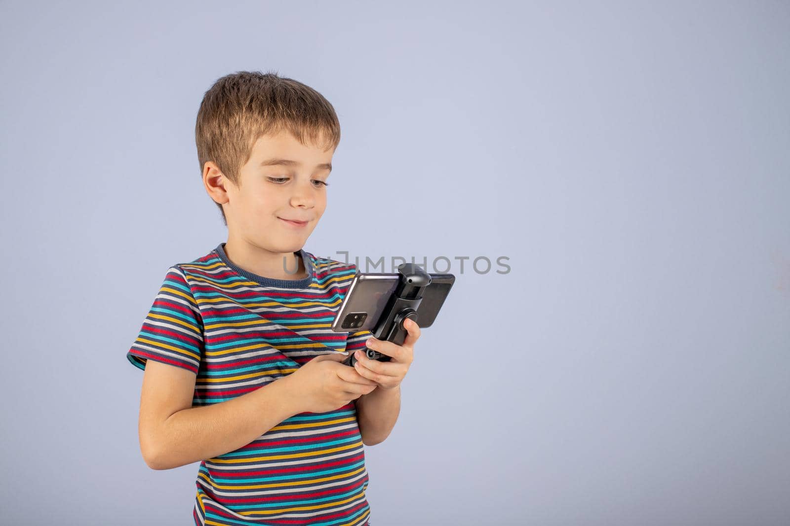 A little boy in a striped T-shirt smiles and looks at the smartphone screen with a holder. On a blue background with a place to copy.