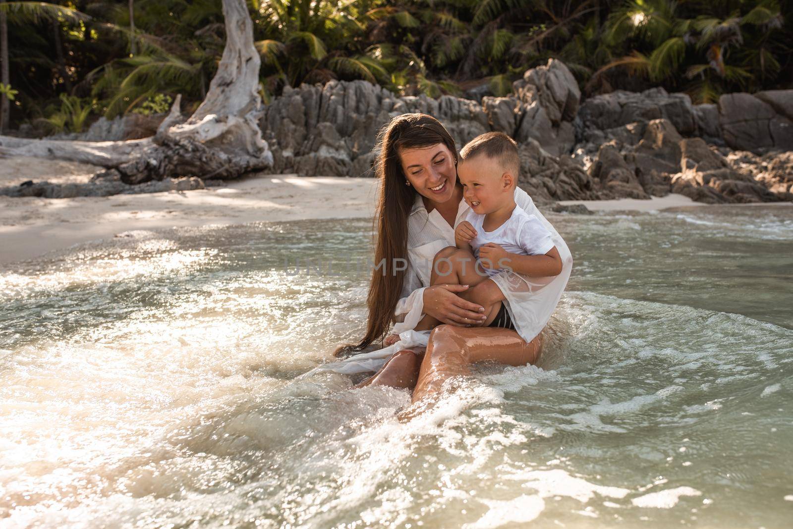 Mother plays with her son in the ocean. Happy smile european family have rest and run on white sand beach. long black chestnut hair mother, blond baby boy. white cotton clothes. boho dress.Thailand.