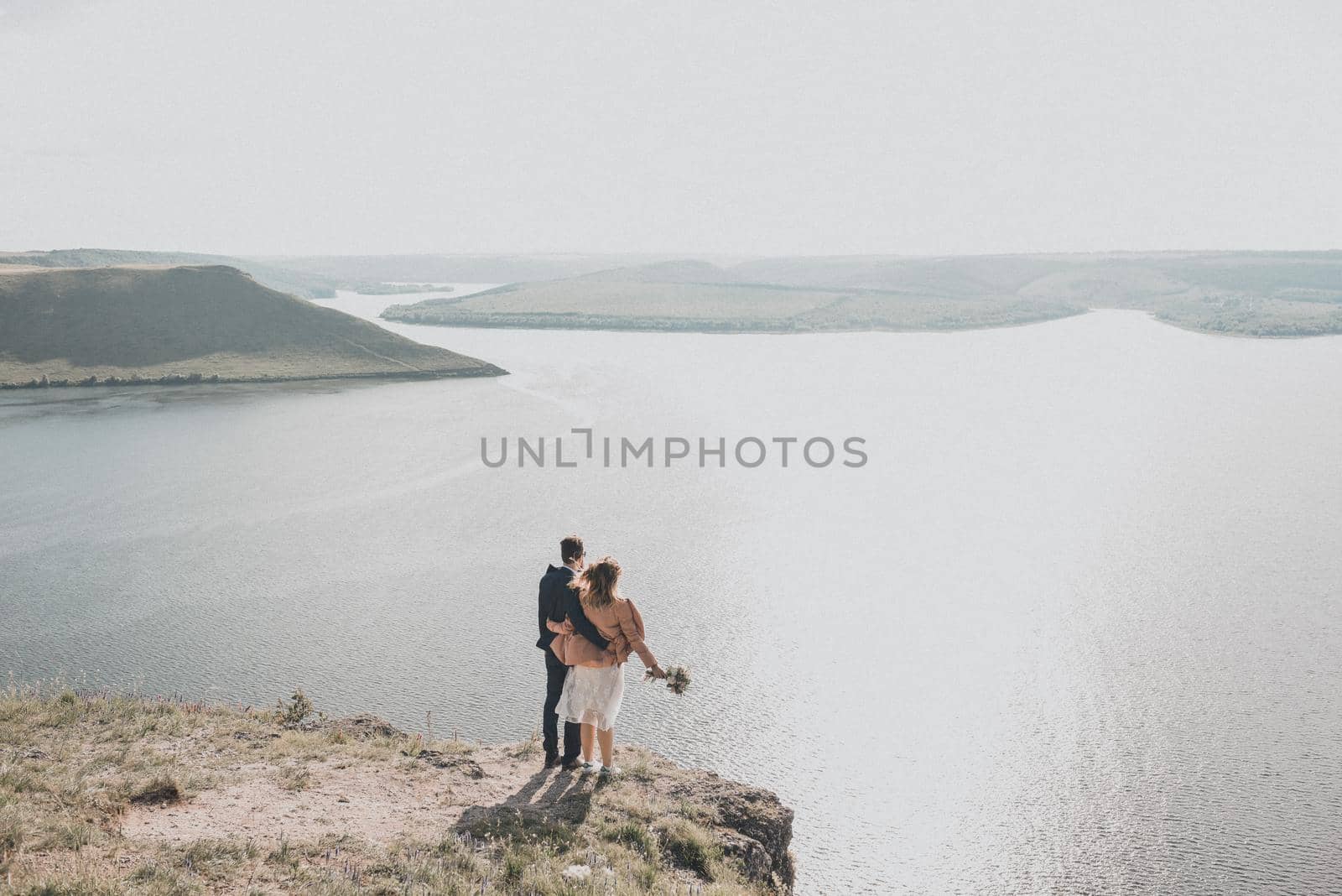 The groom and bride in wedding dress stand on a cliff in front of a large reservoir in distance are islands. Warm sunny summer weather. Spring green grass.A girl in a pink jacket holds a bouquet.