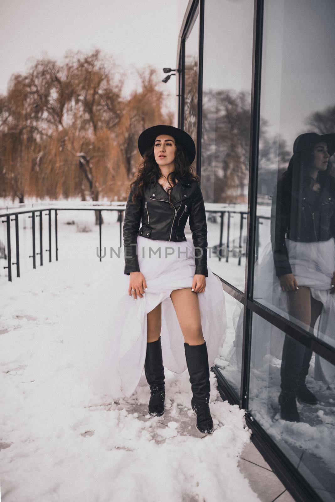 brunette bride woman in wedding white dress, black leather biker jacket and black hat in high boots in winter on the snow. stylish fashion clothing elegant 2021