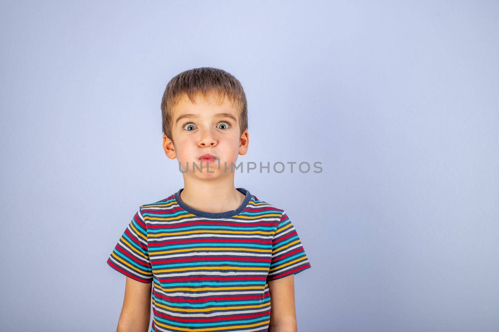 A boy in a striped multicolored T-shirt puffed out his cheeks. Emotions on the face. Blue background. Copy space
