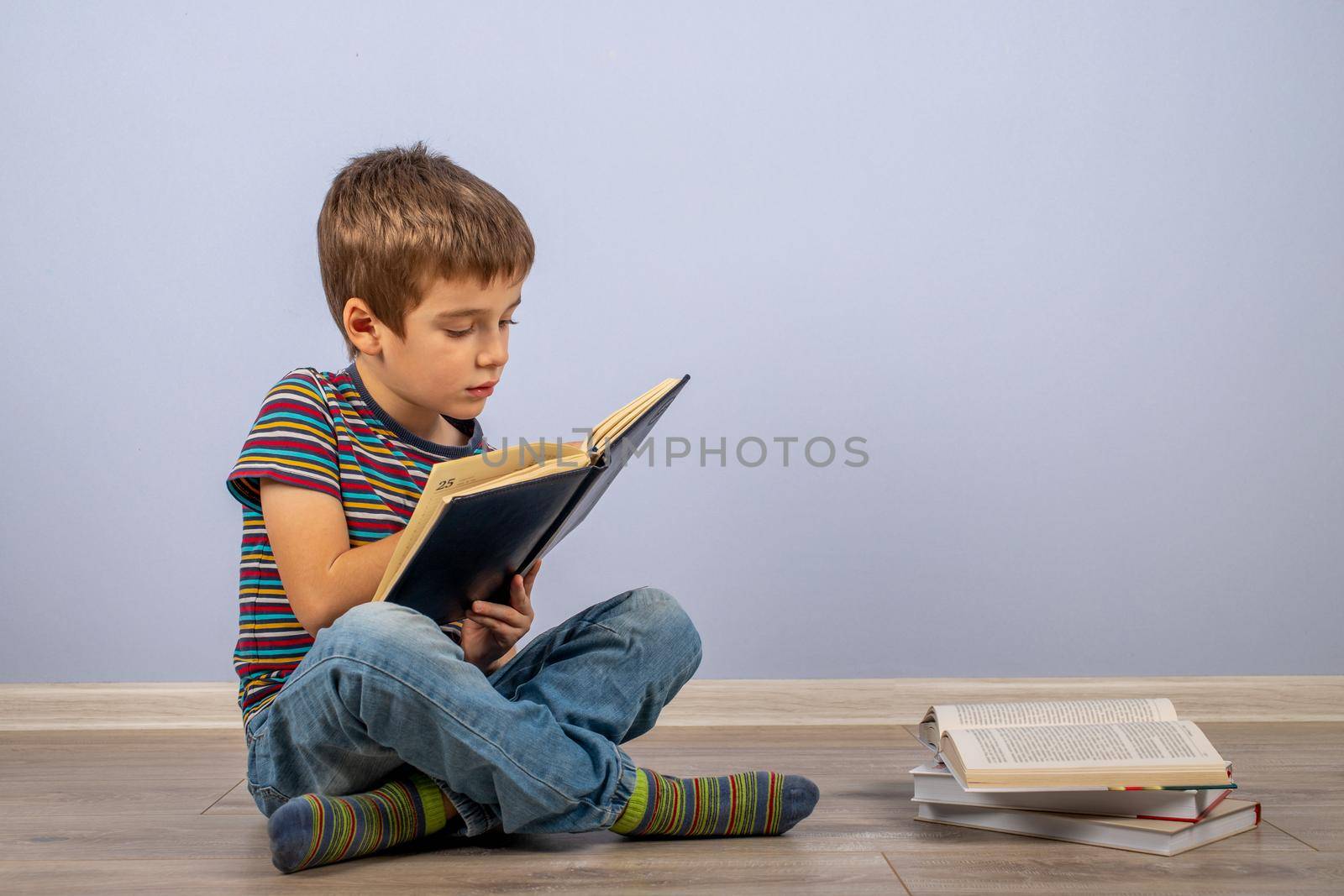 A little boy, studying behind a stack of books and looking at a diary. A male child sitting on the floor on a blue background. by bySergPo
