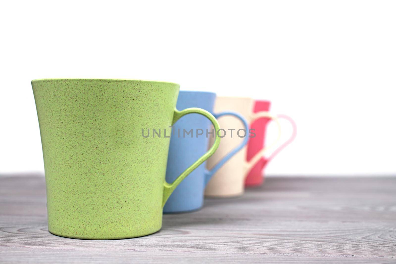 A row of four different-colored plastic mugs on a wooden table on a white background. Dishes made of eco-plastic. The selected focus. by bySergPo