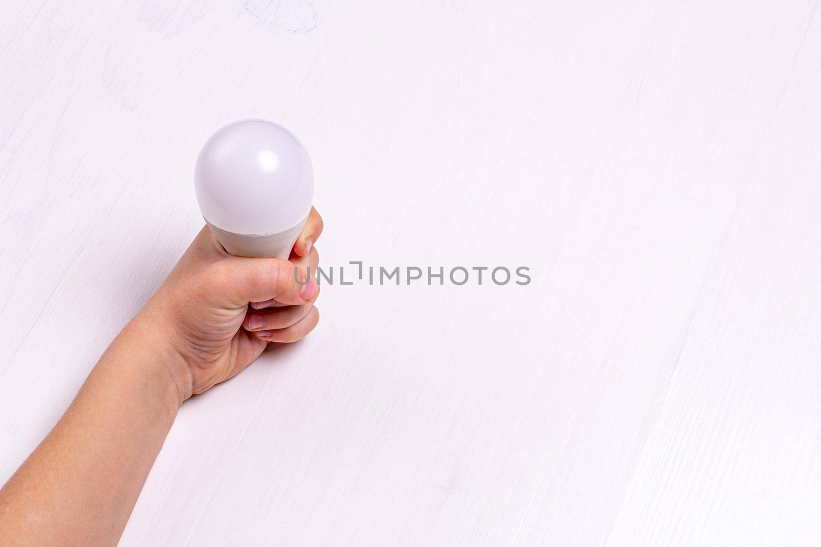 A child's hand holds an LED light bulb against a light background by bySergPo