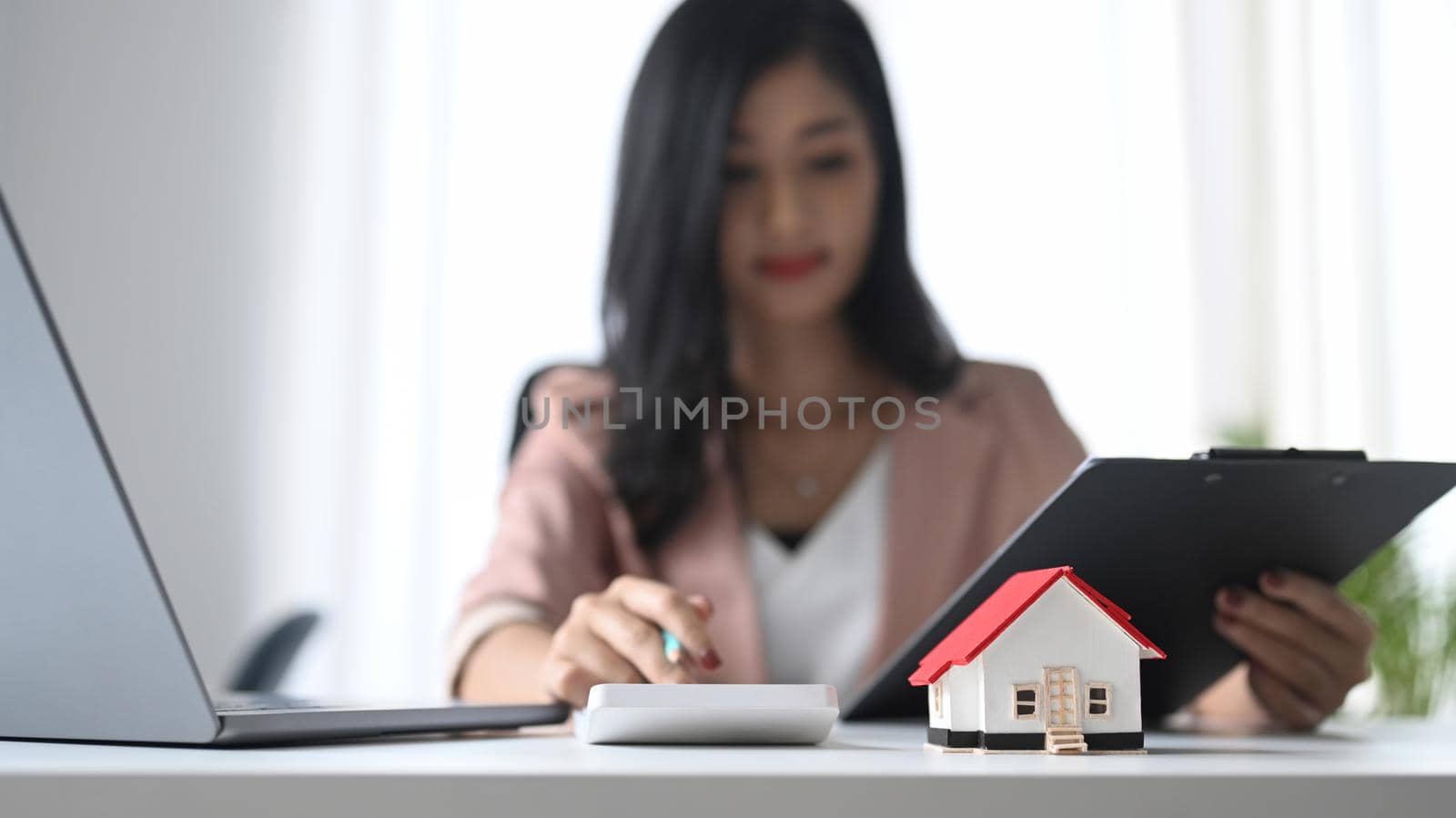 Small house model on white table and professional financial advisor is using calculator.