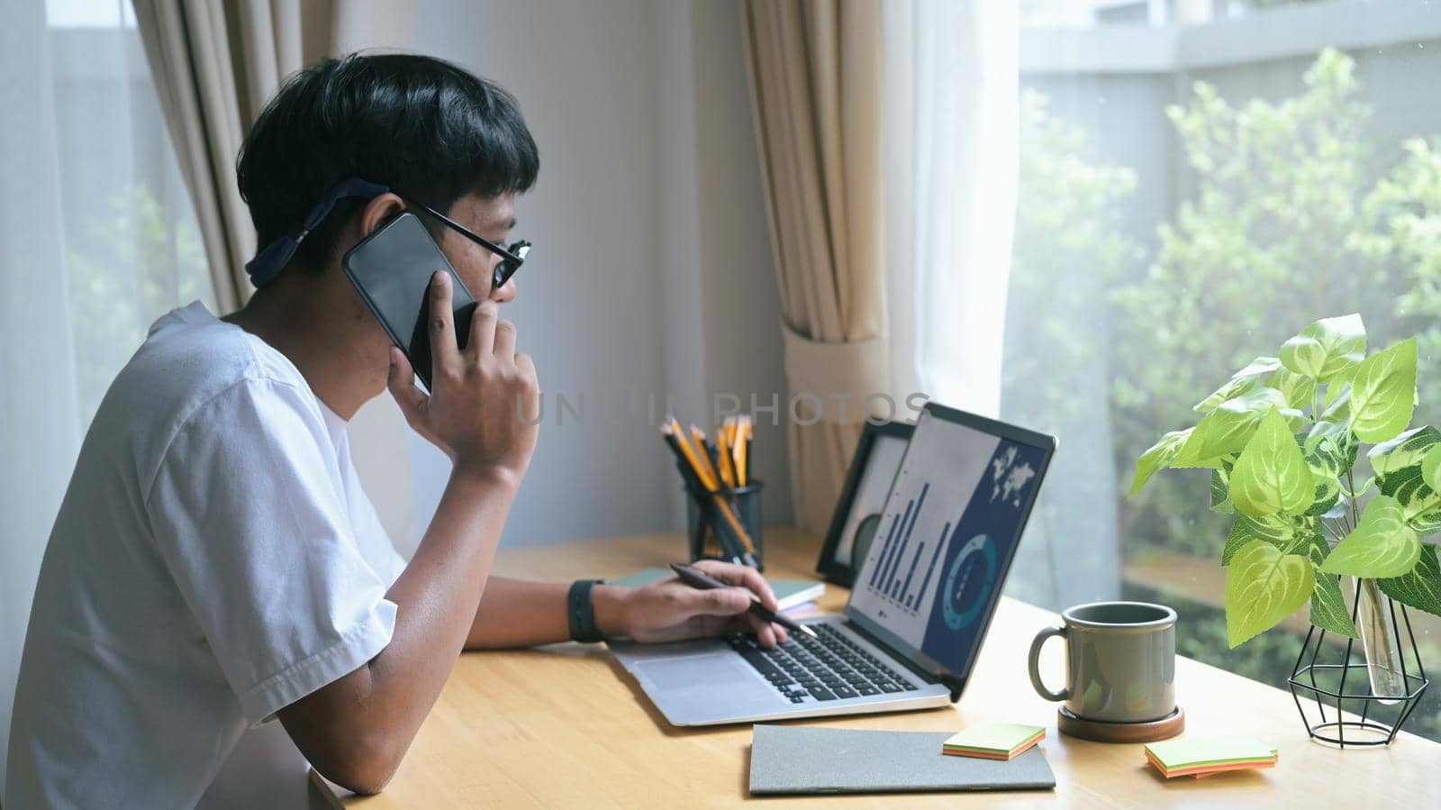 Young man having cell phone conversation and working with computer laptop a home.