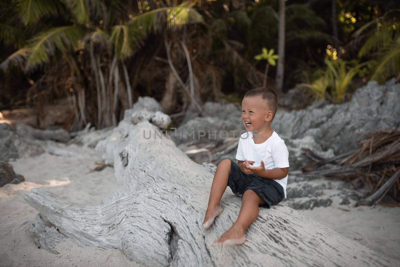 Happy smile european blond baby boy have rest and sit on white sand beach. white cotton clothes. t-short.Thailand. palms. rocks