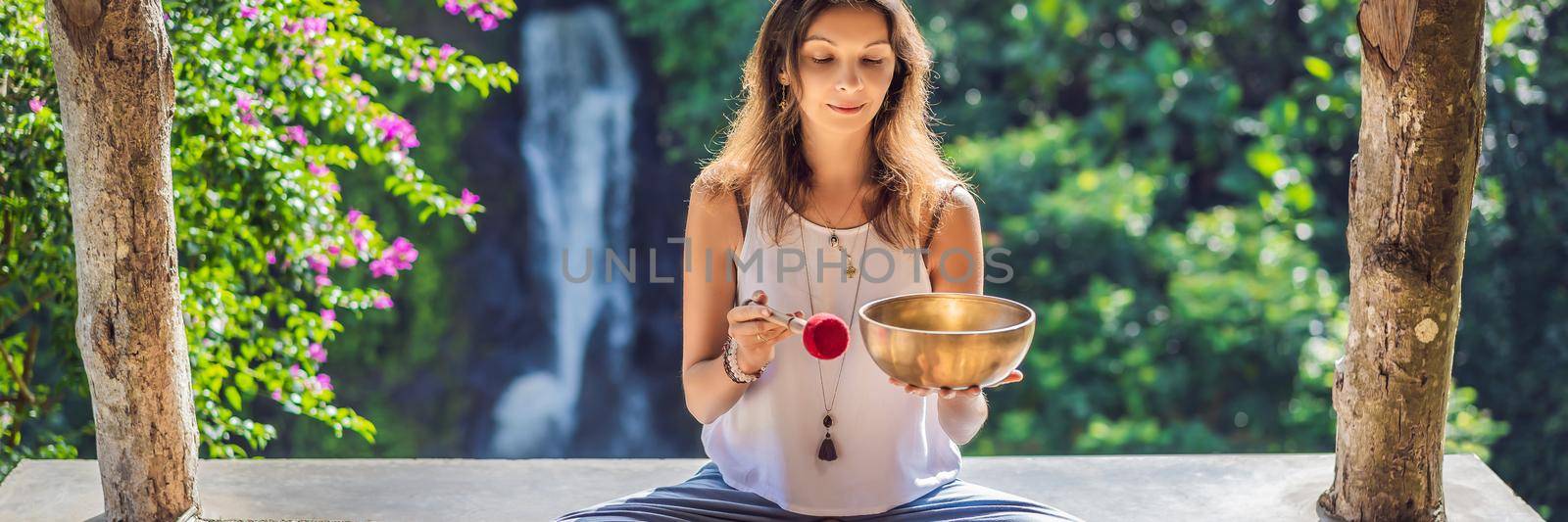 Woman playing on Tibetan singing bowl while sitting on yoga mat against a waterfall. Vintage tonned. Beautiful girl with mala beads meditating. BANNER, LONG FORMAT