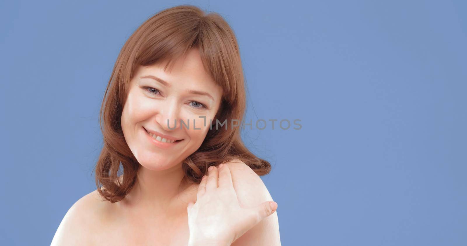 Mature brown-haired female applies skin care cream by touching face with hand, close up. Woman applying anti-age cream on her skin isolated on blue background. Spa and wellness concept.