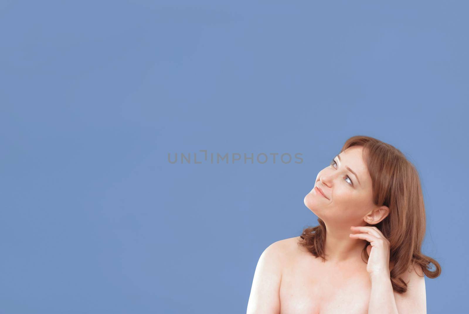 Naked woman applying anti-age cream on her skin isolated on color background. Pretty mature female applies skin care product by touching face with hand. Close up. Spa and wellness concept. Template.