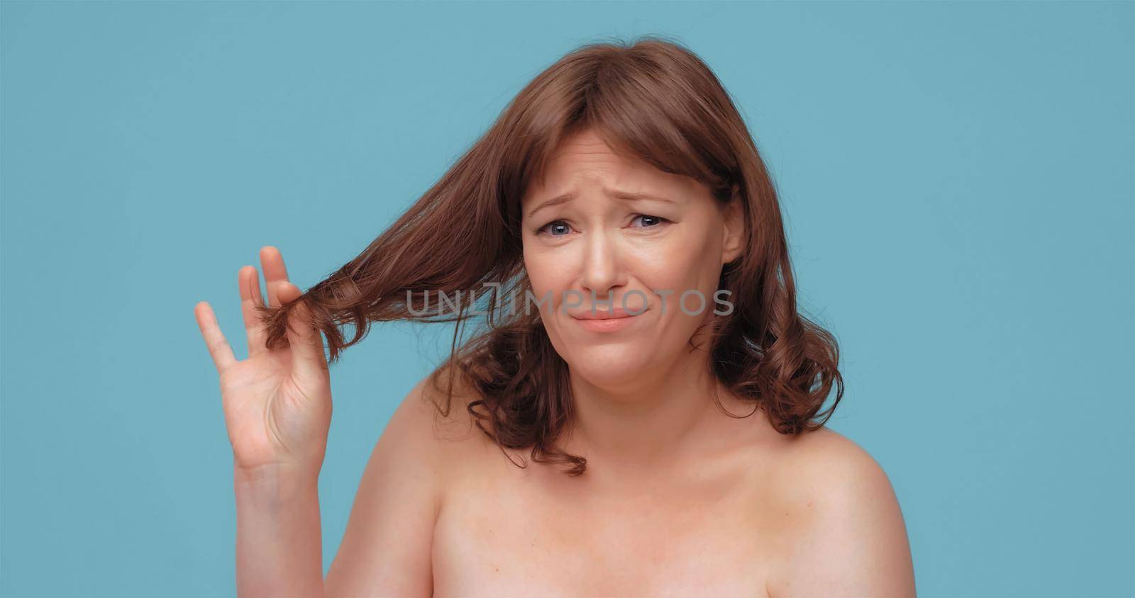 Unhappy beautiful woman holds of hair ends looking at camera. Pretty nude mature woman on color back. Hair care concept by LipikStockMedia