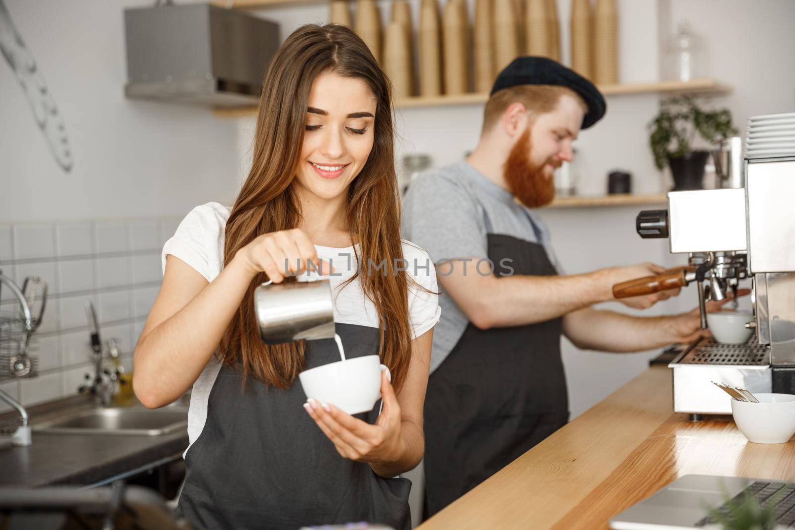 Coffee Business Concept - close-up lady barista in apron preparing and pouring milk into hot cup while standing at cafe. by Benzoix