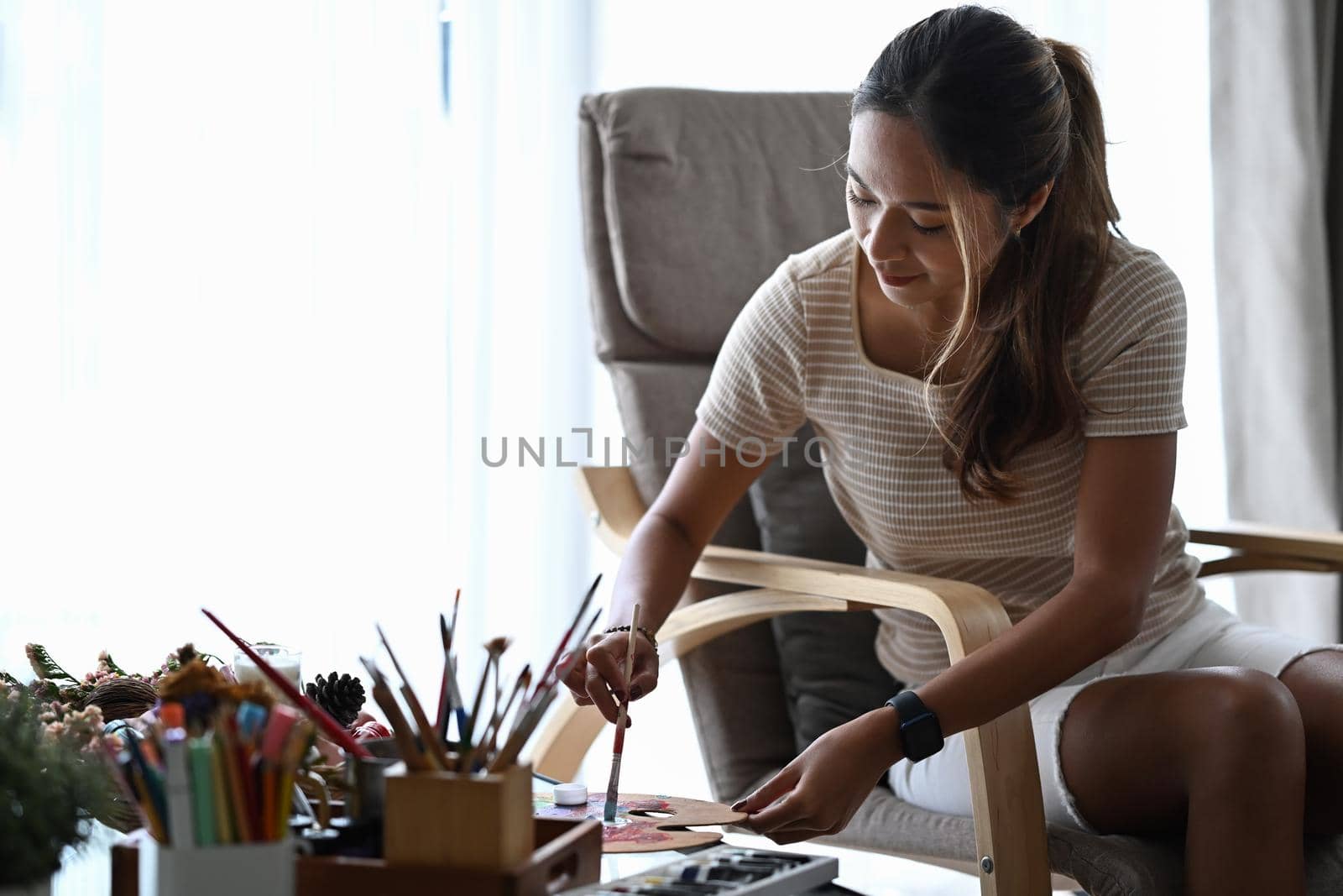 Beautiful woman sitting on armchair in living room and mixing color oil paint on palette.