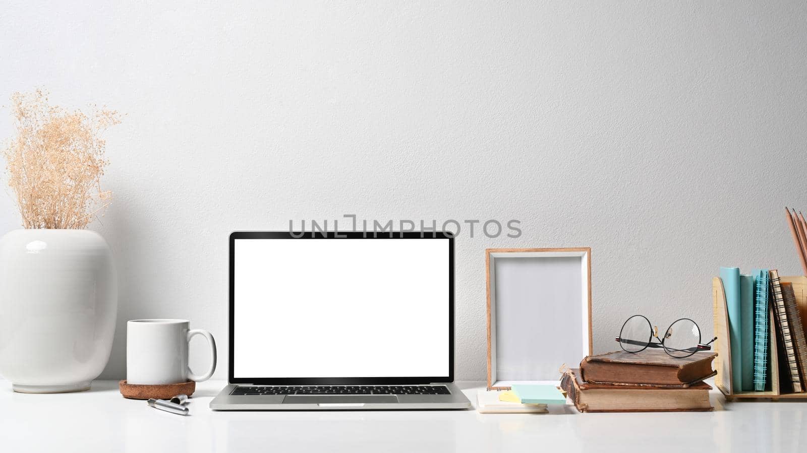 Comfortable workplace with laptop computer, picture frame, flower pot, coffee cup and stationery on white table.