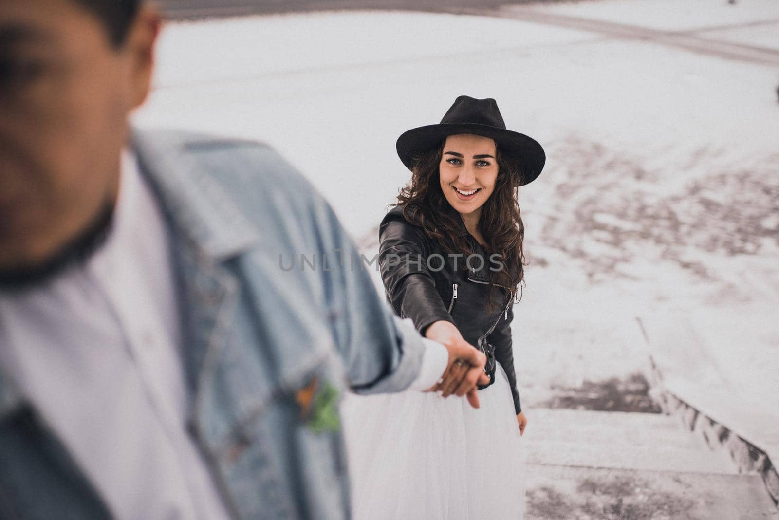 Mexican Hispanic man in oversized denim jacket and woman in hat leather biker jacket kissing hugging laughing walking.groom bride wedding love couple in winter snow.fashionable stylish party wedding