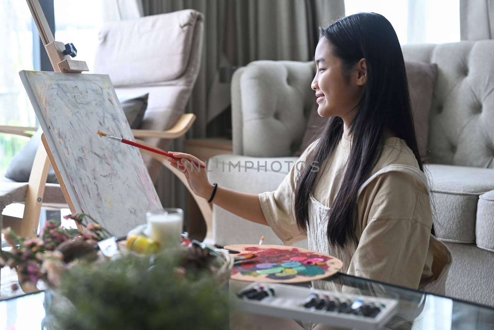 Smiling asian girl painting picture on easel in living room.