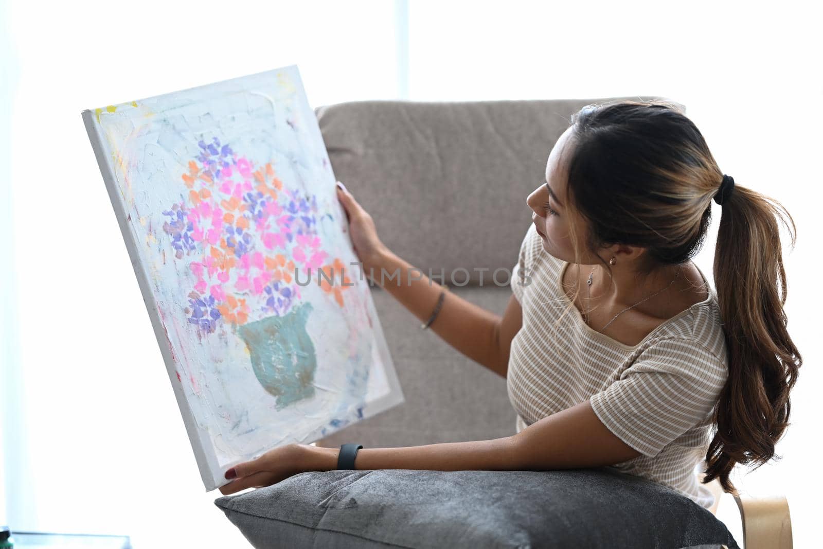 Pleased young woman sitting on armchair in living room and looking at her artwork with admiration.