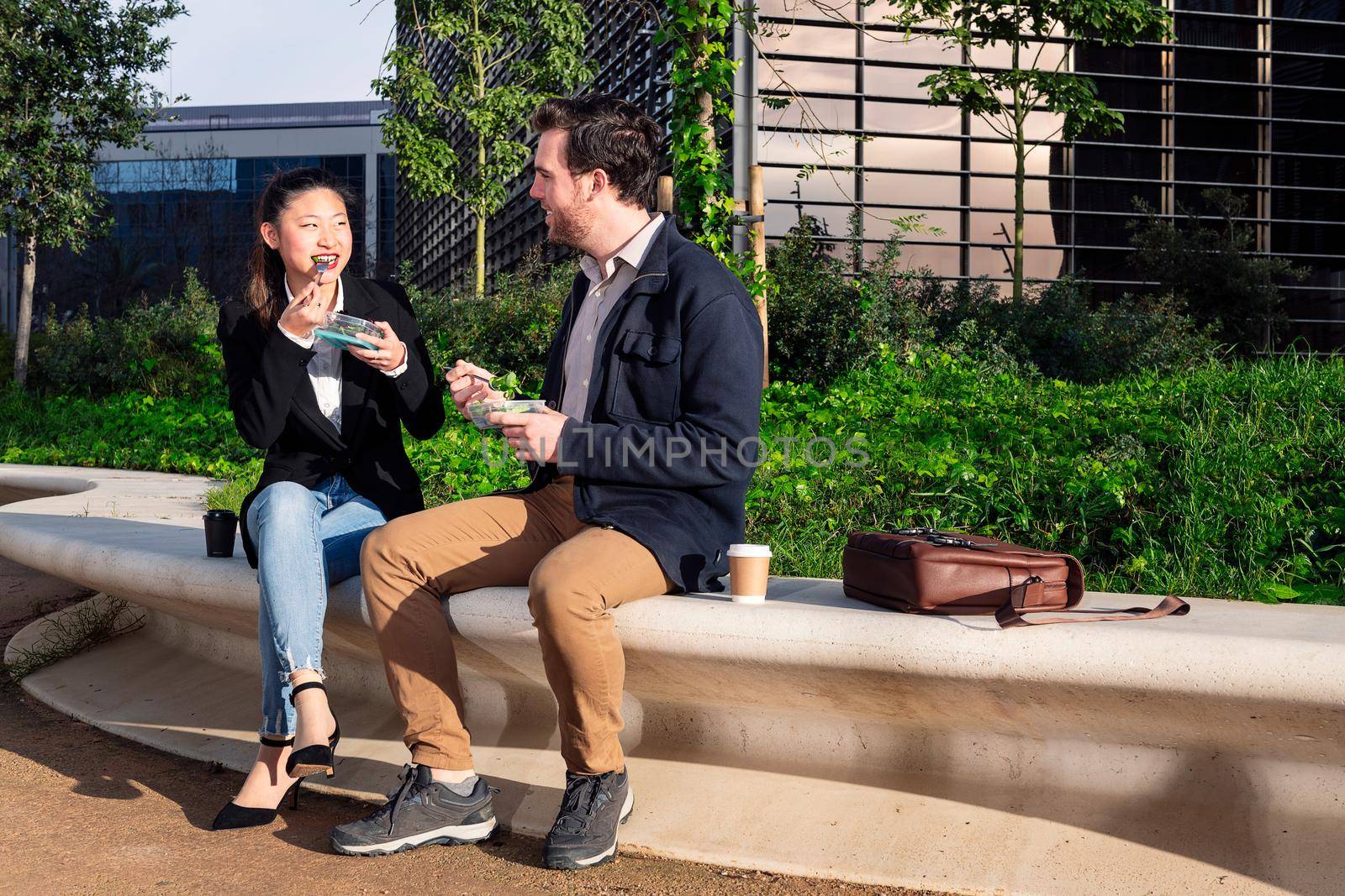 asian woman talking and eating a salad with her caucasian partner sitting in a park next to the office, concept of healthy fast food at work, copy space for text