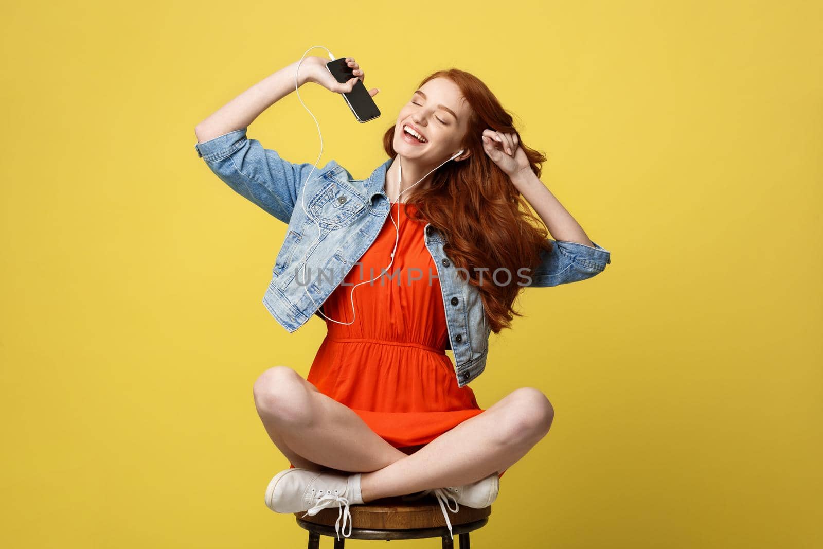 Lifestyle Concept - Happy pretty red hair woman in earphones listening to music and singing while sitting on chair and holding mobile phone. Isolated on bright yellow background by Benzoix