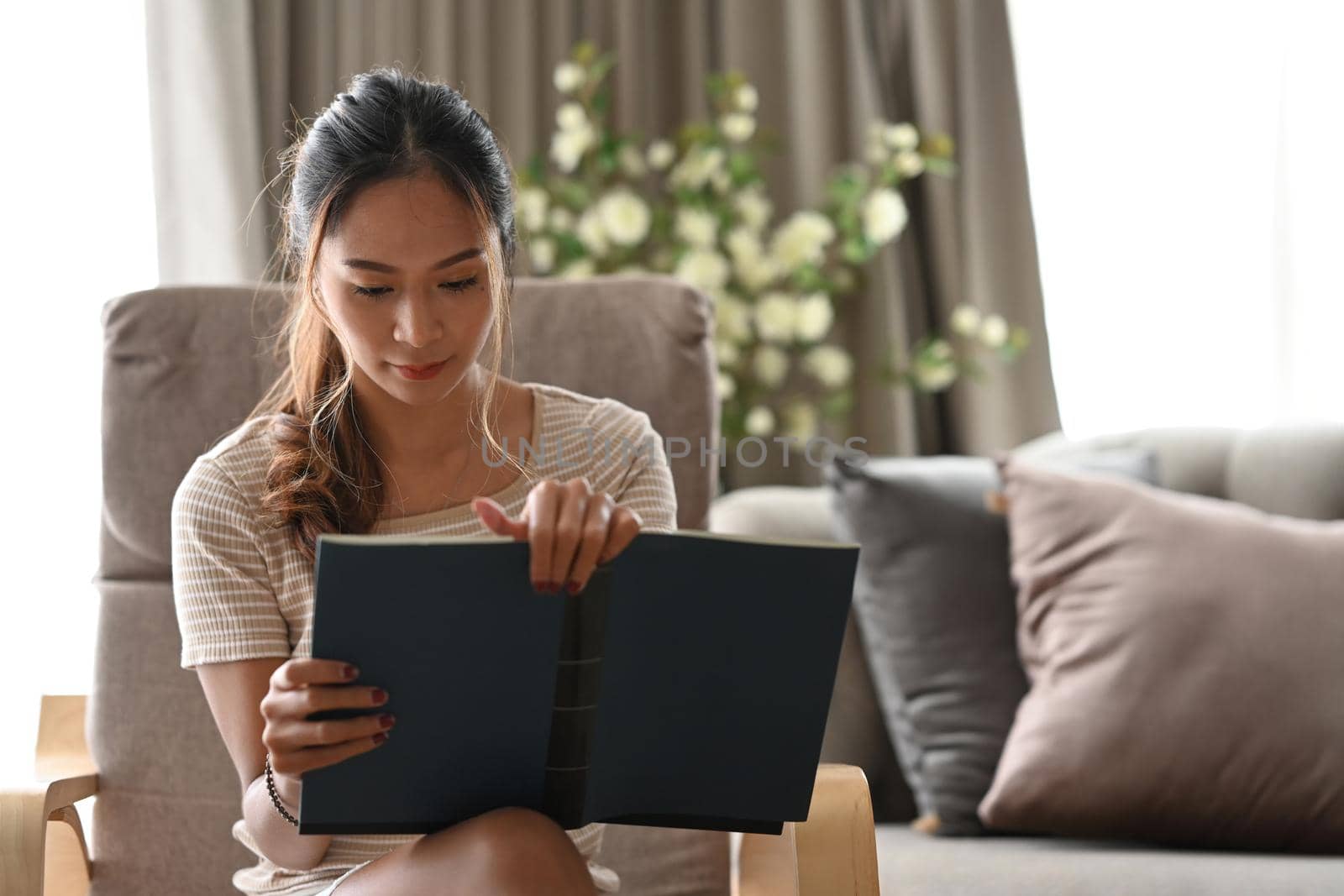 Peaceful asian woman reading book while sitting on beige armchair in her apartment.