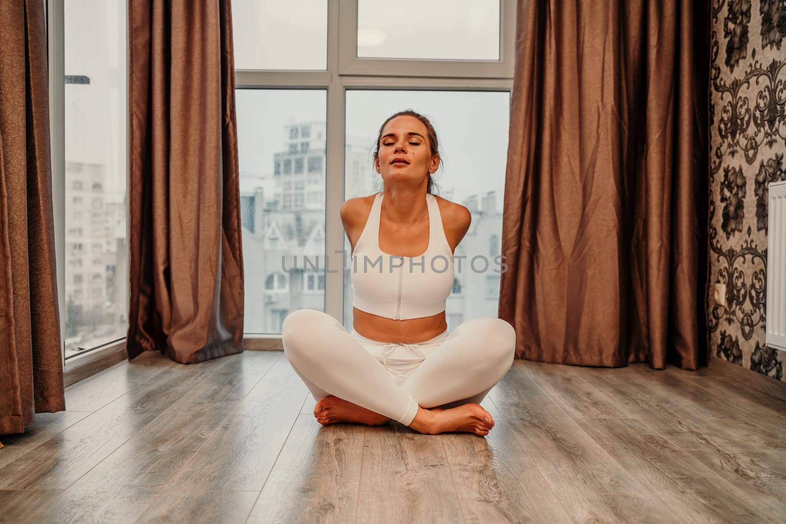 Middle aged athletic attractive woman practicing yoga. Works out at home or in a yoga studio, sportswear, white pants and a full-length top indoors. Healthy lifestyle concept by Matiunina
