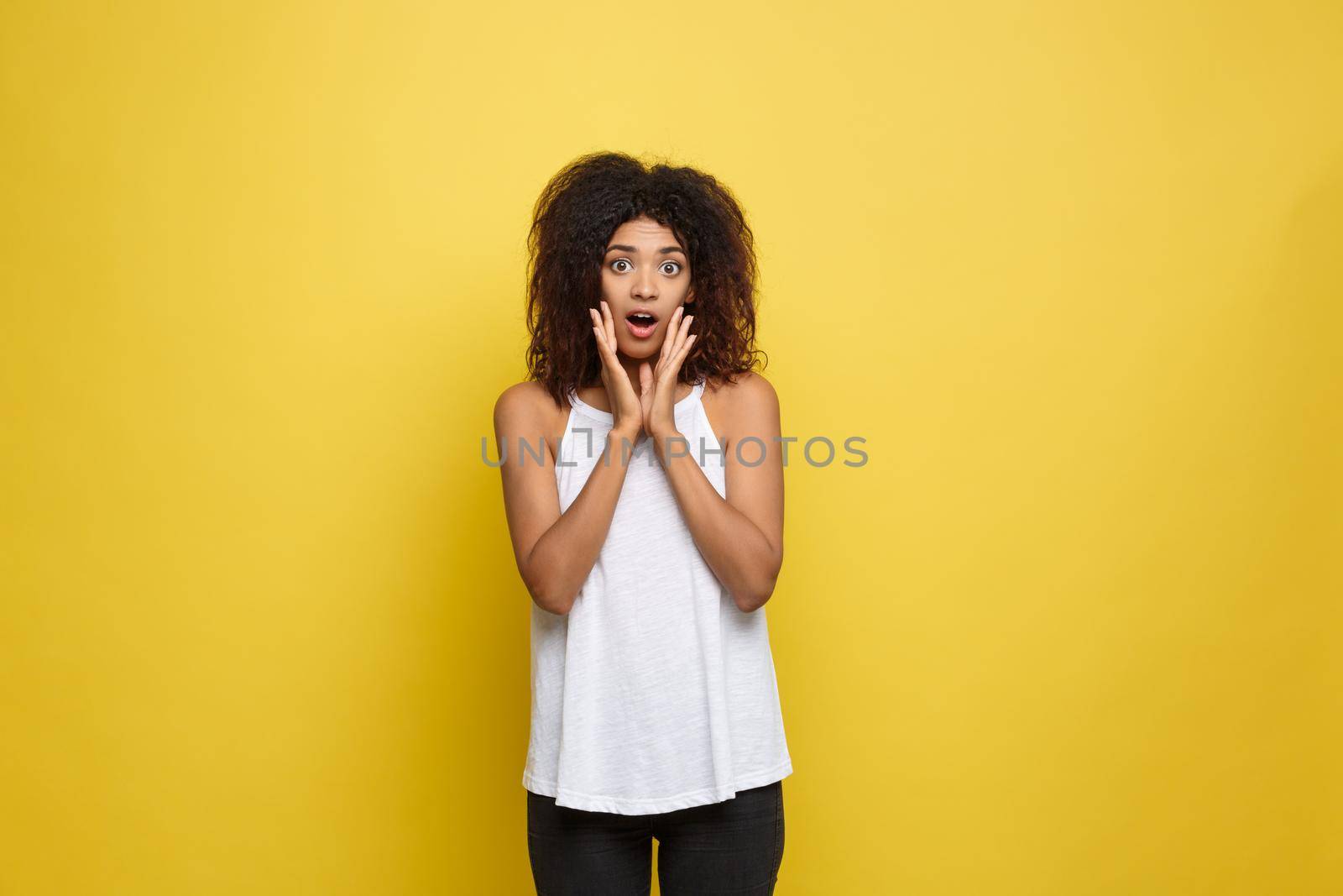 Smiling beautiful young African American woman in white T-shirt posing with hands on chin. Studio shot on Yellow background. Copy Space. by Benzoix