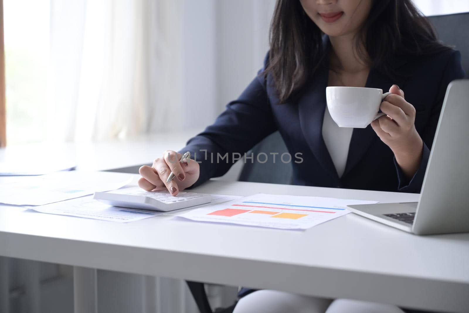 Female financial advisor drinking coffee and using calculator at office desk.