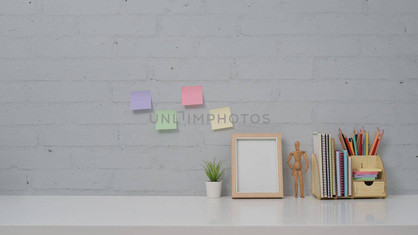 Home office desk with picture frame, potted plant, stationery and colorful sticky notes on brick wall background.
