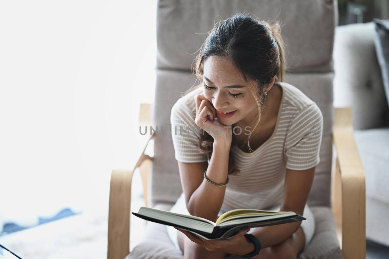 Satisfied young woman reading book on armchair while spending leisure time at home.