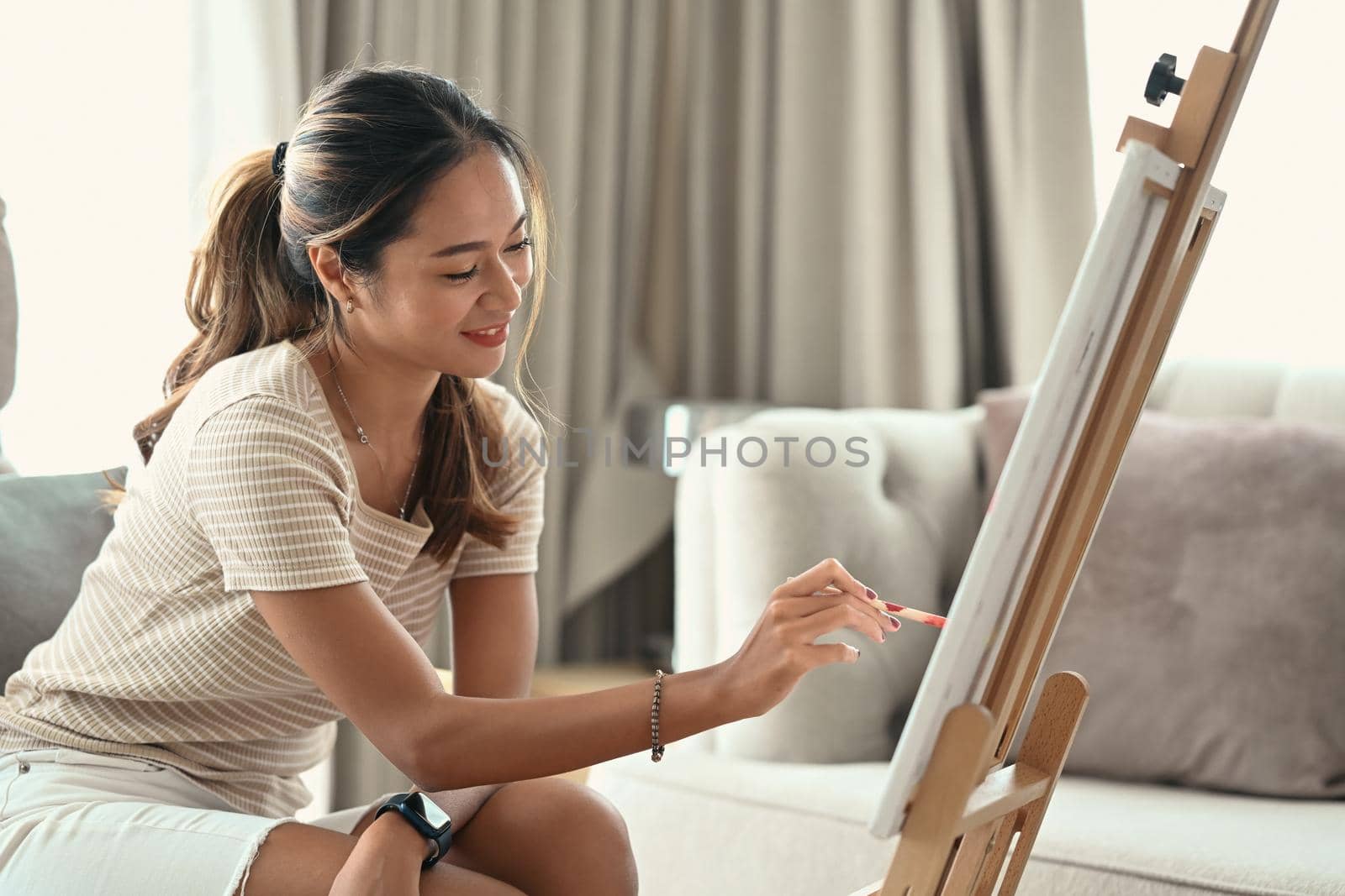 Charming young woman painting picture on canvas with oil paints at home.