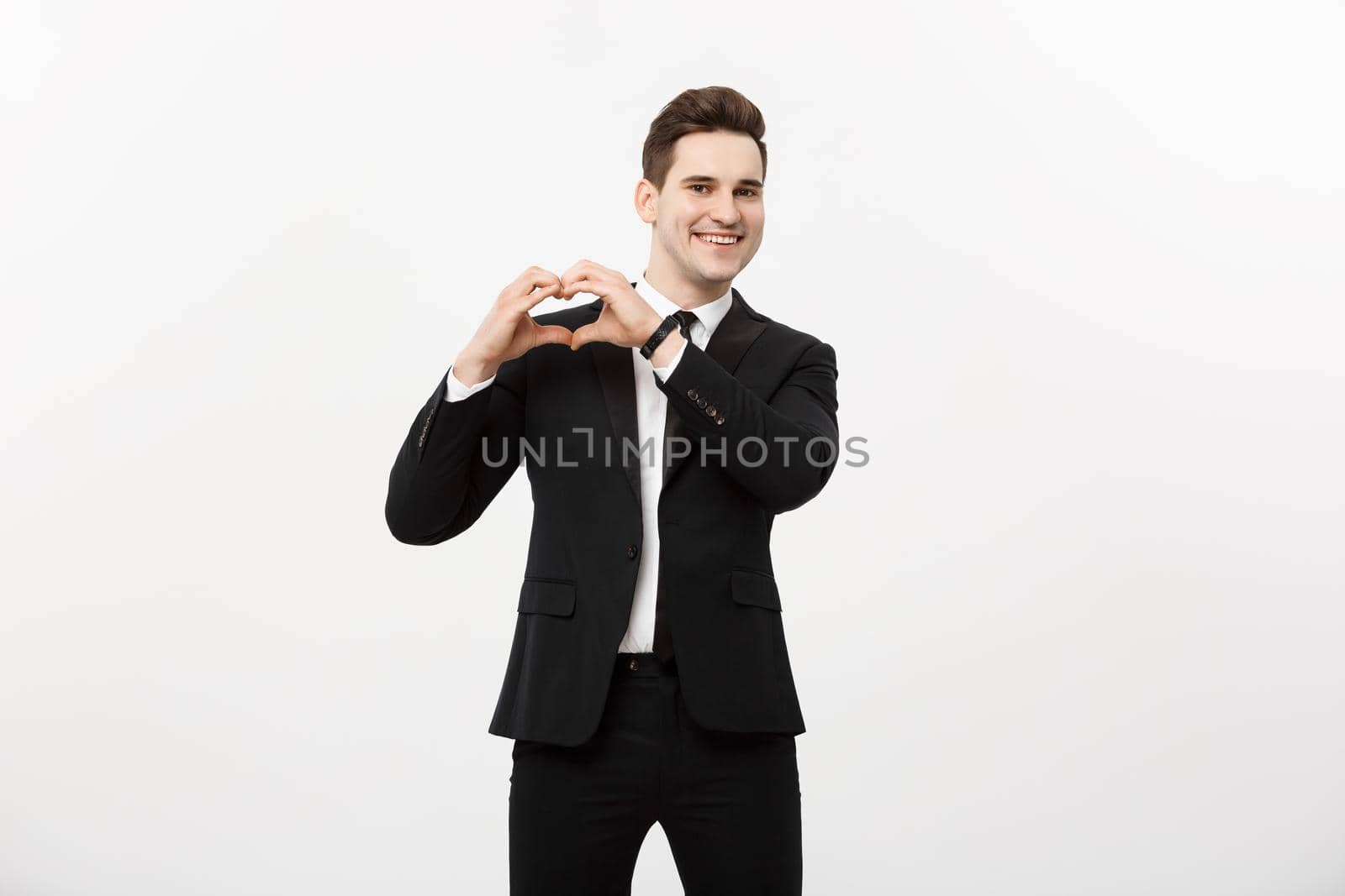 Business Concept: Portrait of charming attractive businessman holding hands in heart gesture and lifting eyebrows while smiling, isolated over white grey background. by Benzoix