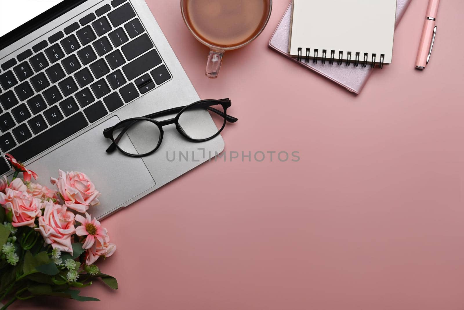 Home office desk with blank notebook, laptop computer and pink roses bouquet on pink background. Flat lay.