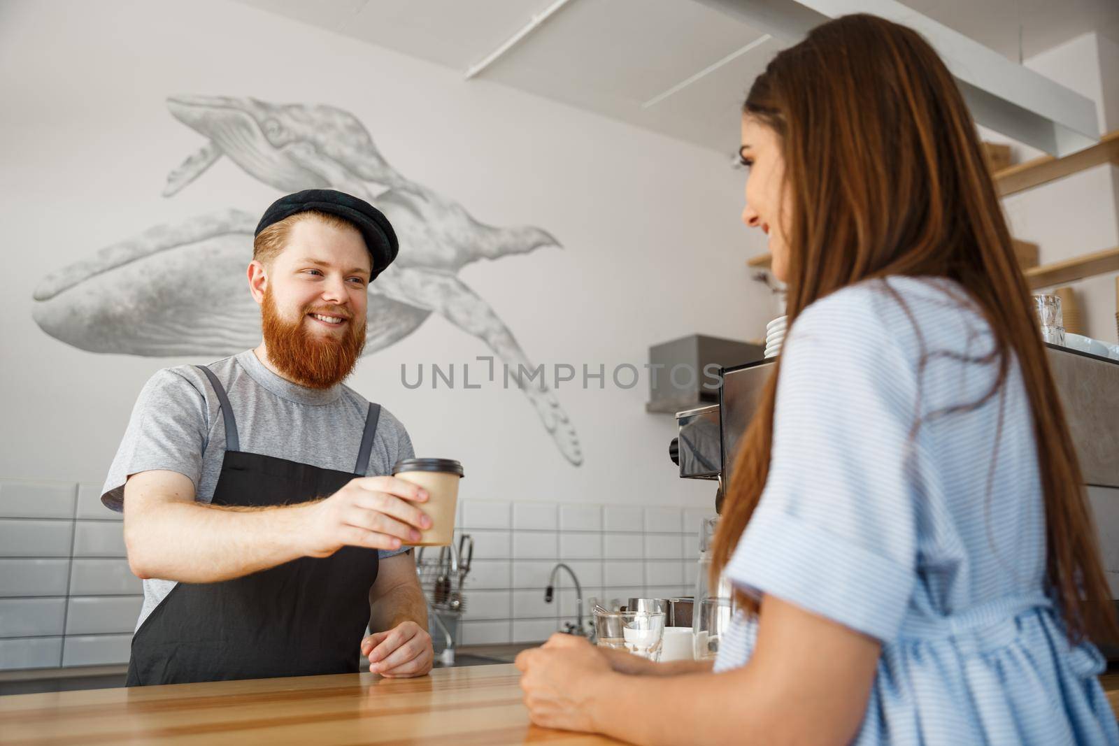 Coffee Business Concept - young beared handsome barista talking and giving take away hot coffee for beautiful caucasian customer in modern coffee shop.