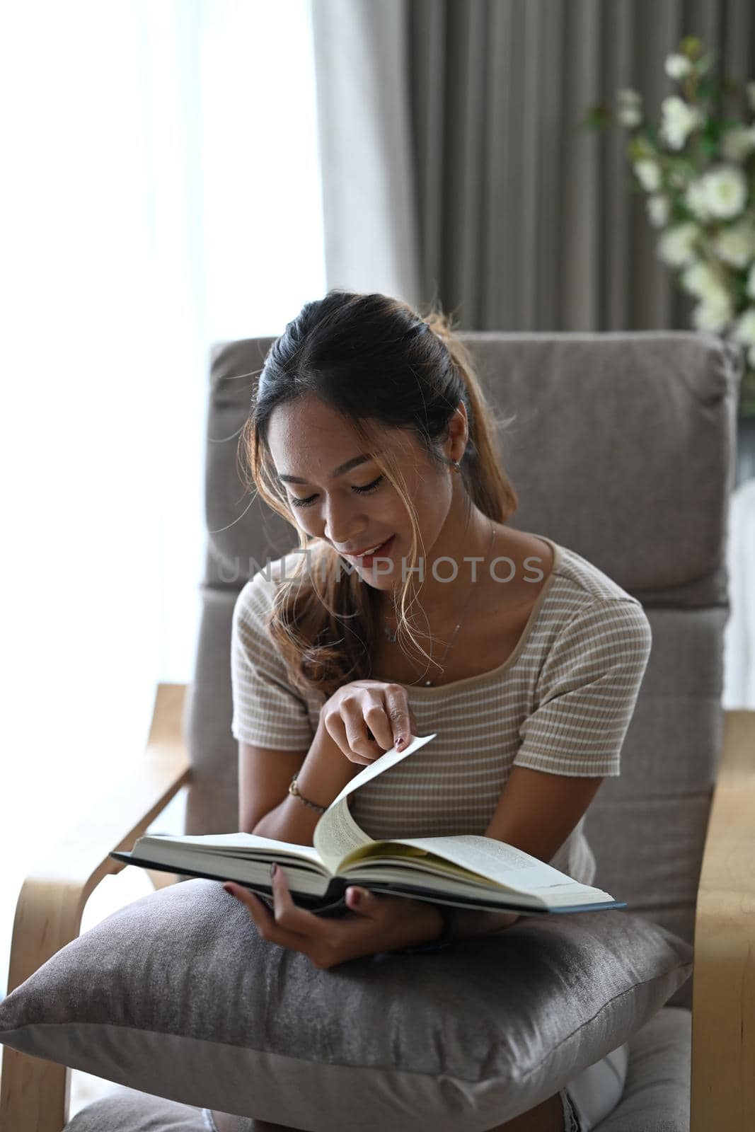 Happy young woman resting on armchair in living room and reading her favorite novel book.