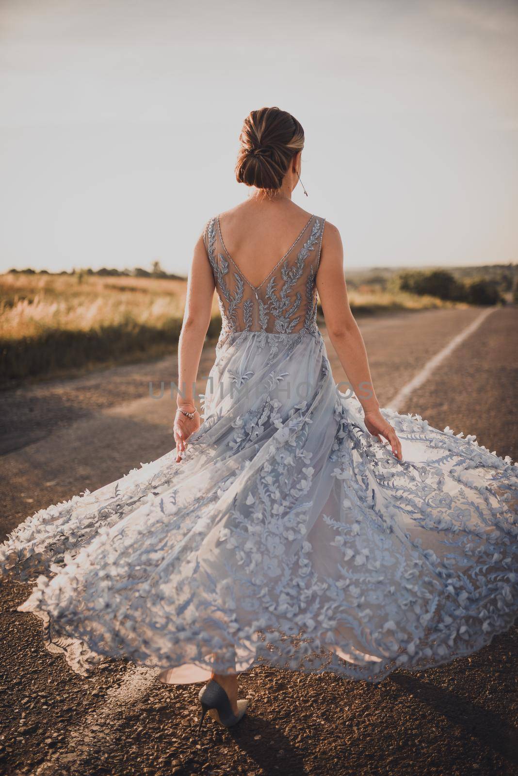 a young girl walks along an asphalt road in a gray blue long dress at sunset. back and arms open. Near the road tall yellow grass grows. The background is blurred.