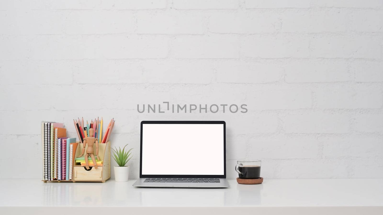 Front view computer laptop, coffee cup and stationery on white table against brick wall. by prathanchorruangsak