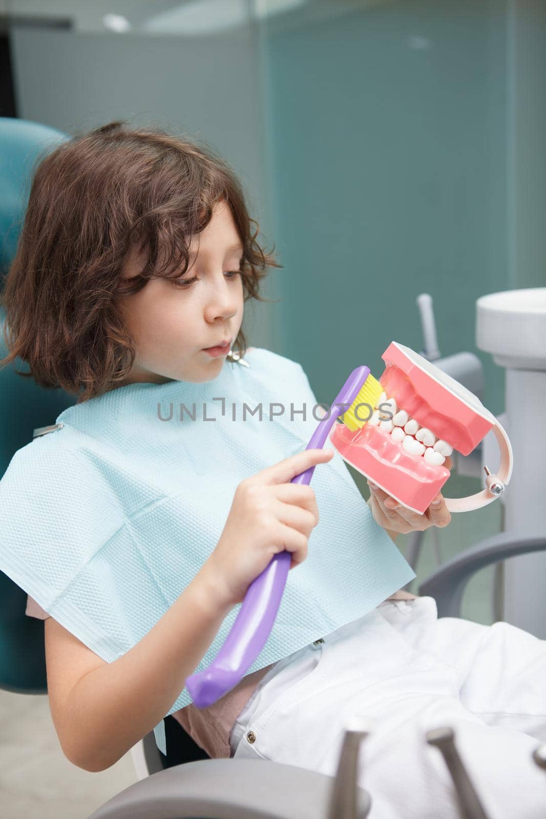 Vertical shot of a cute young boy brushing jaw model with toothbrush at dental clinic
