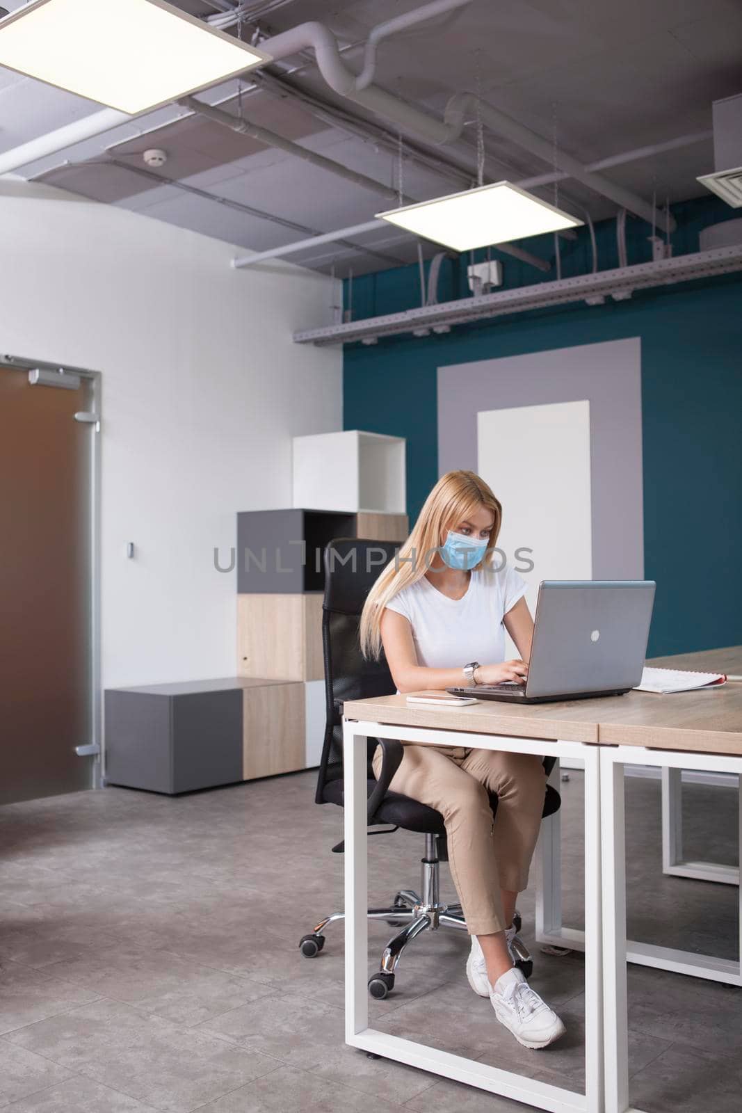 Vertical full length shot of a businesswoman wearing protective face mask working on her laptop at open space office
