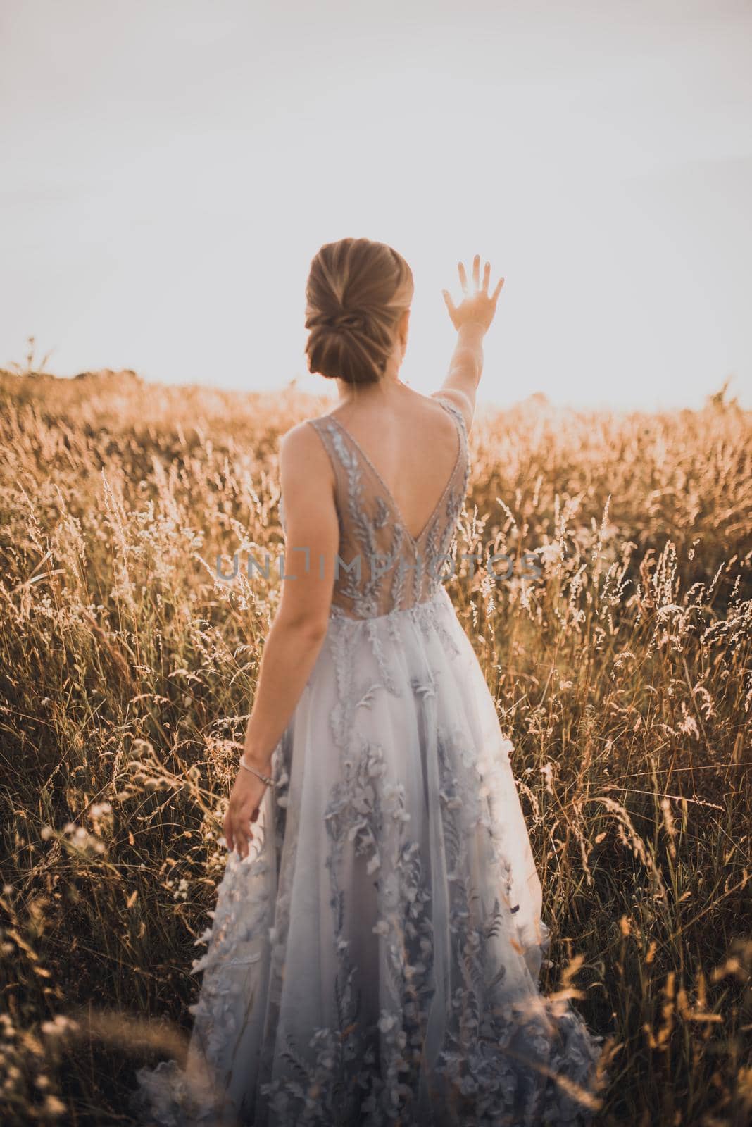 The young beautiful girl in a gray dress with an open back raised her hands up and closes the rays of the sun. against the background of blurred tall yellow grass at sunset.