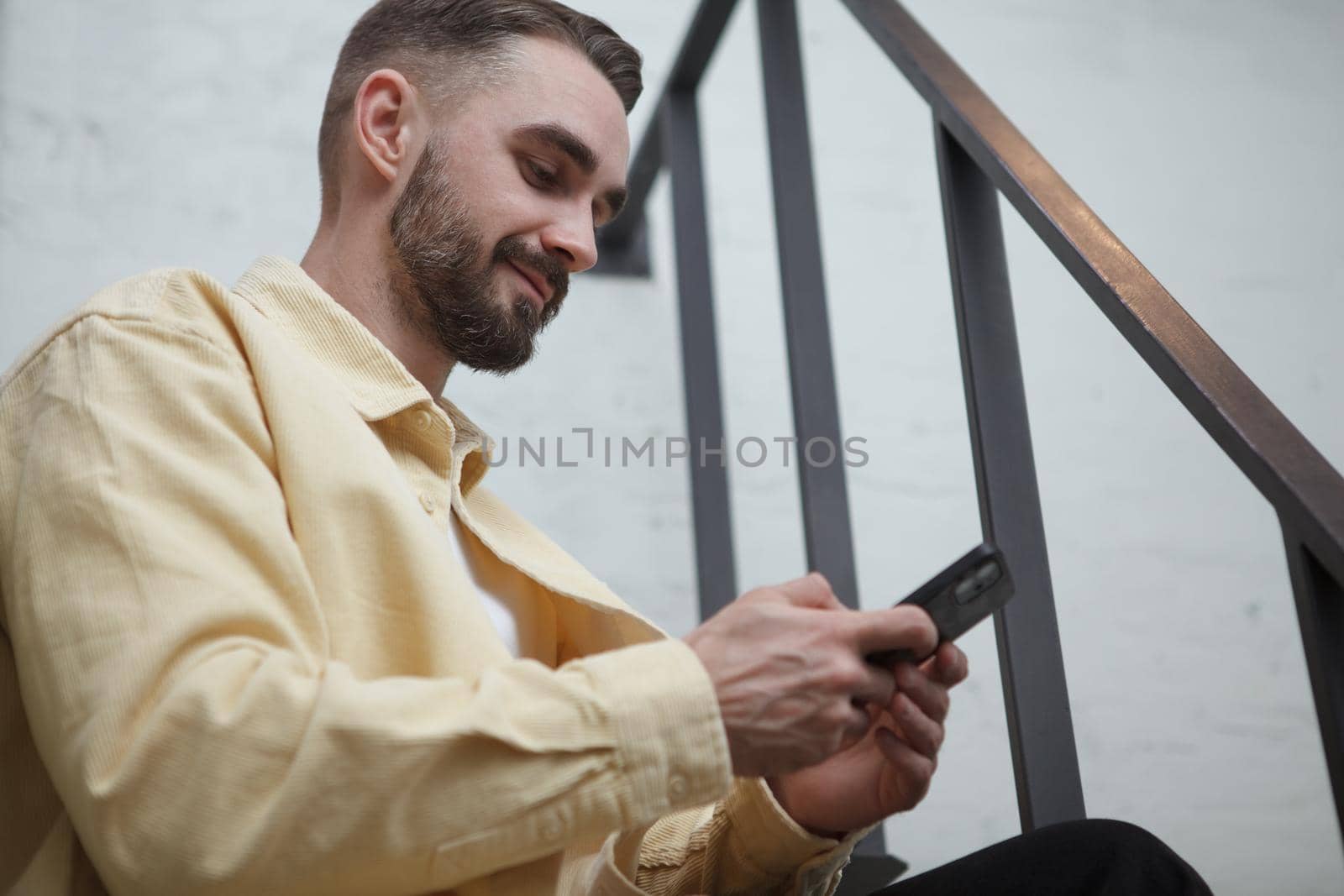 Handsome bearded man smiling, using smart phone app, sitting on the stairs indoors