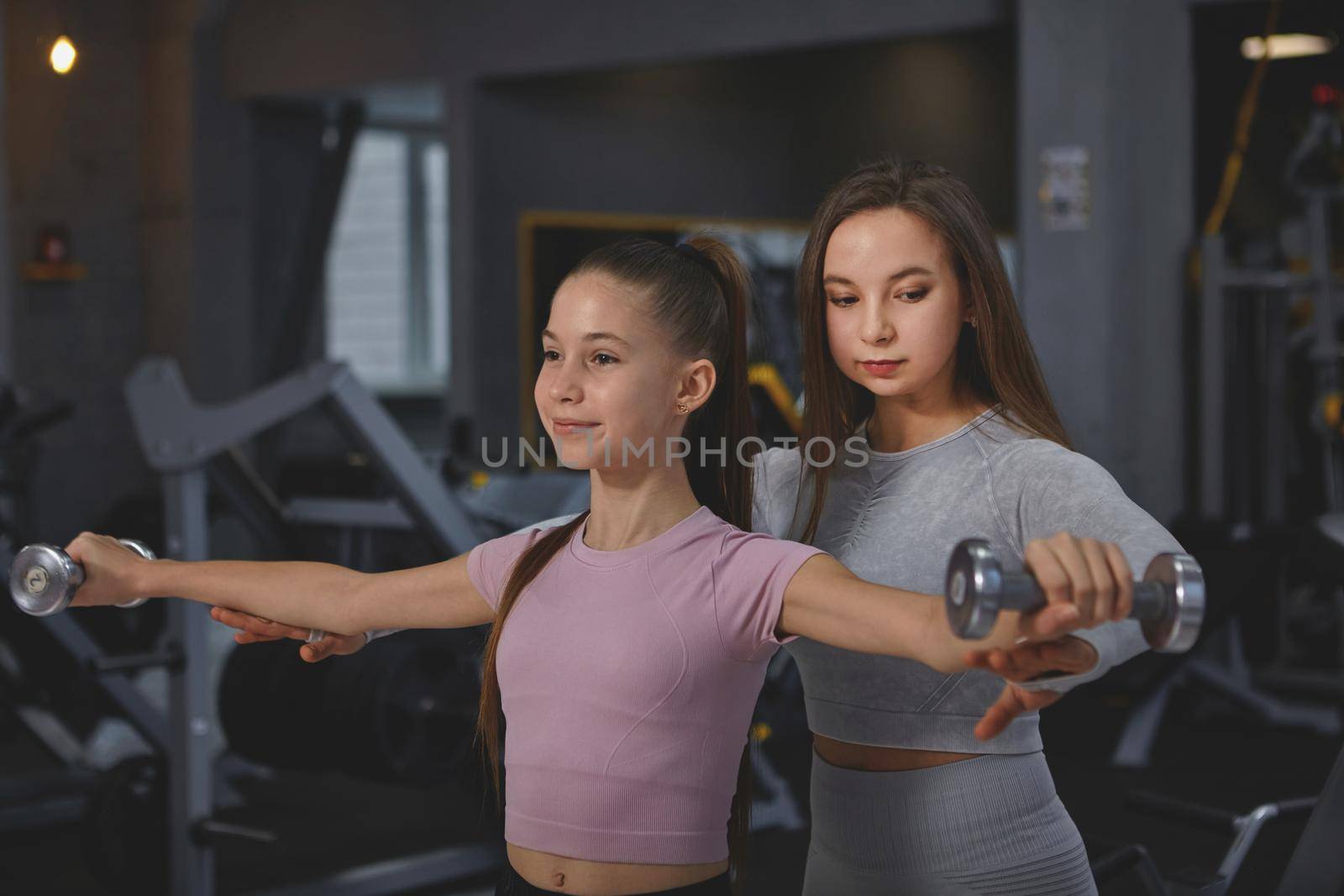 Professional sportswoman supervising teenage girl during gym workout, doing dumbbells exercise