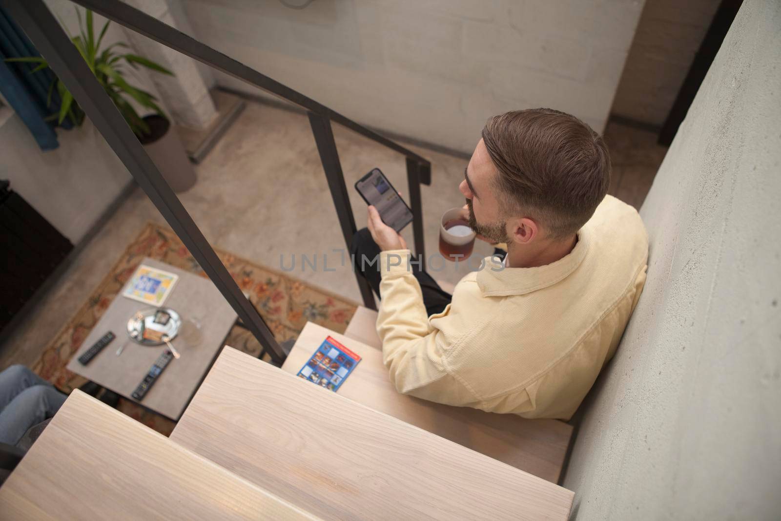Top view shot of a man having tea while texting on his smart phone, sitting on staircase indoors