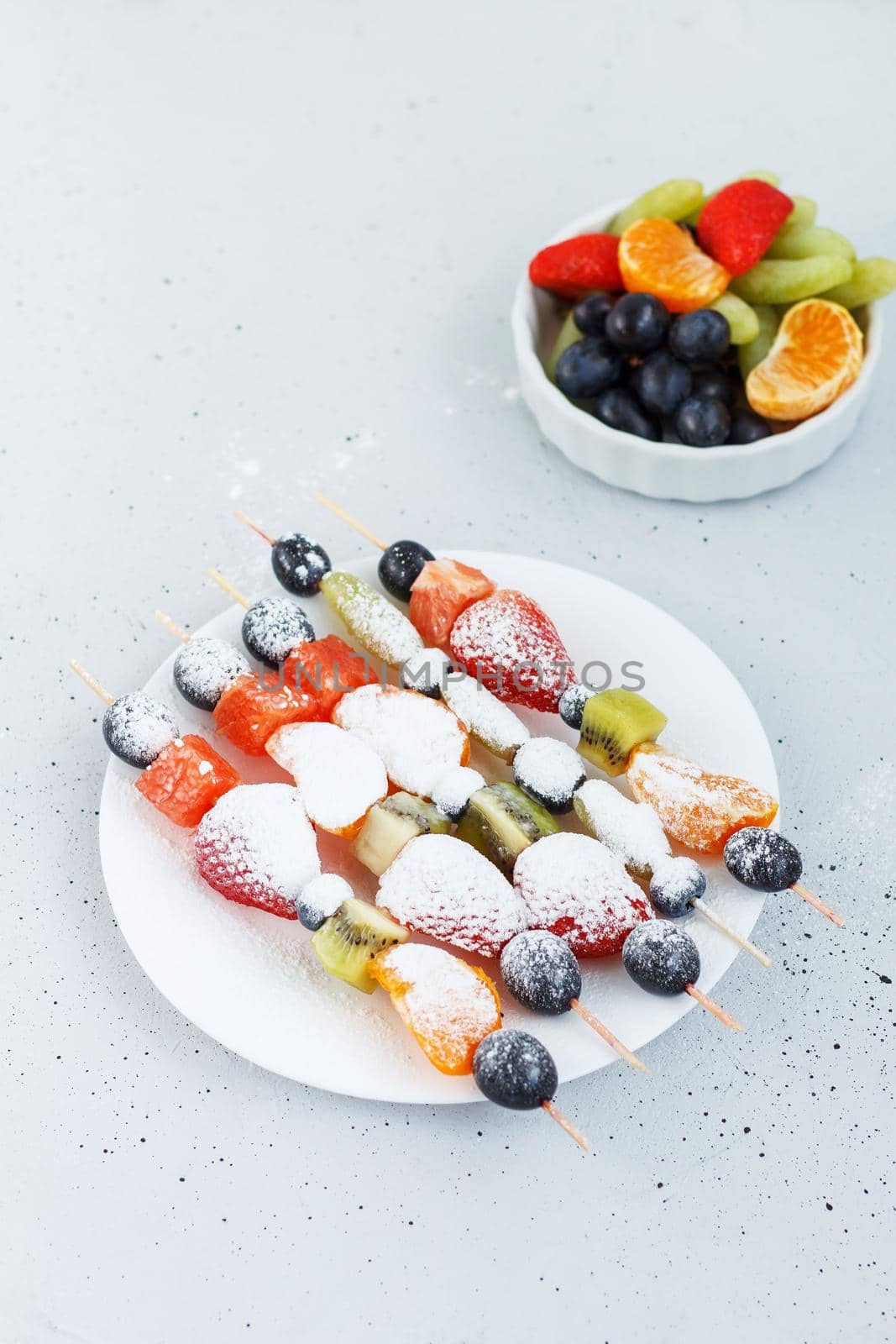 Fresh fruit skewers on a plate sprinkled with powder for a healthy snack on a gray background. Vertical photo by lara29