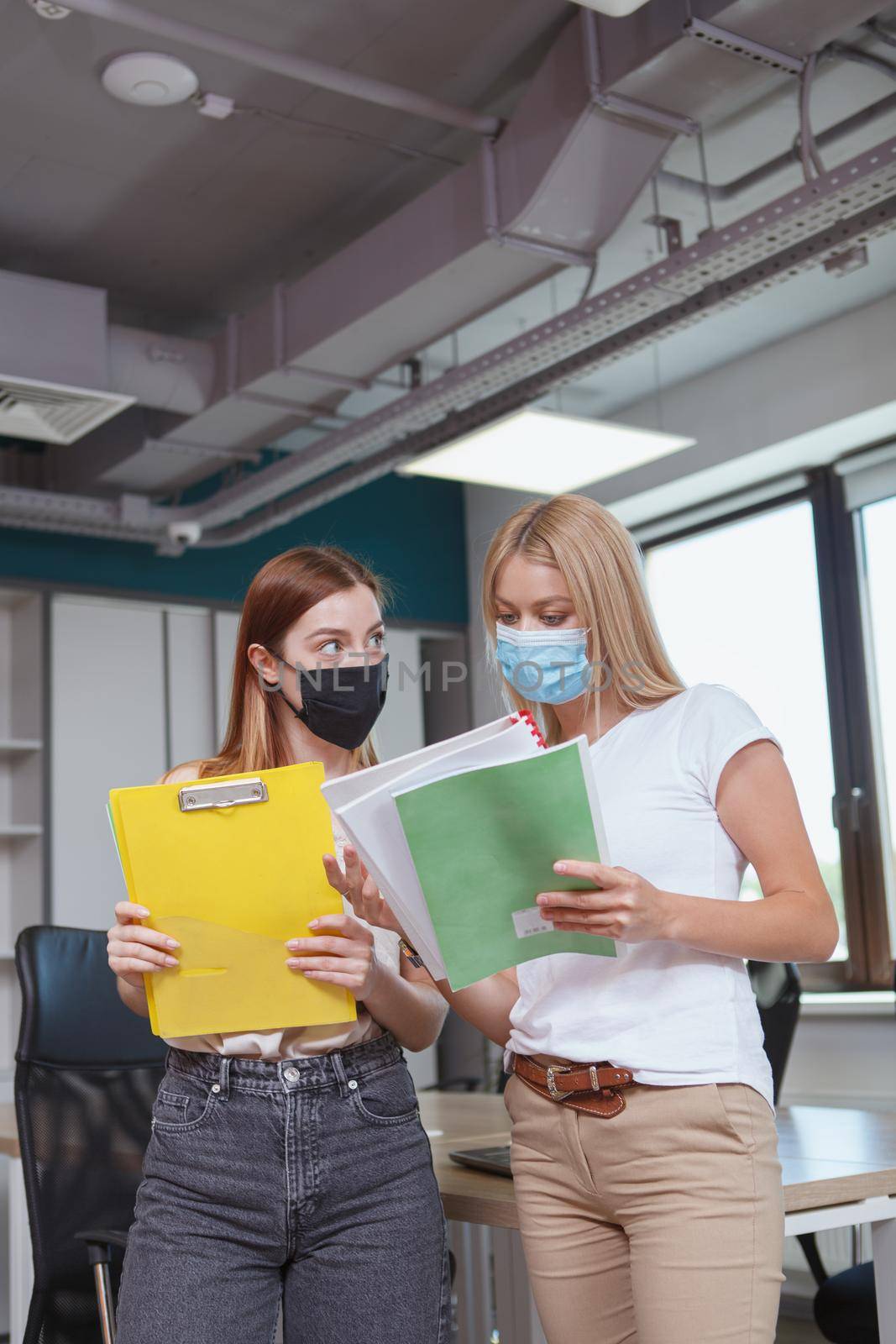 Vertical shot of two female colleagues wearing face masks, working at the office during covid pandemic