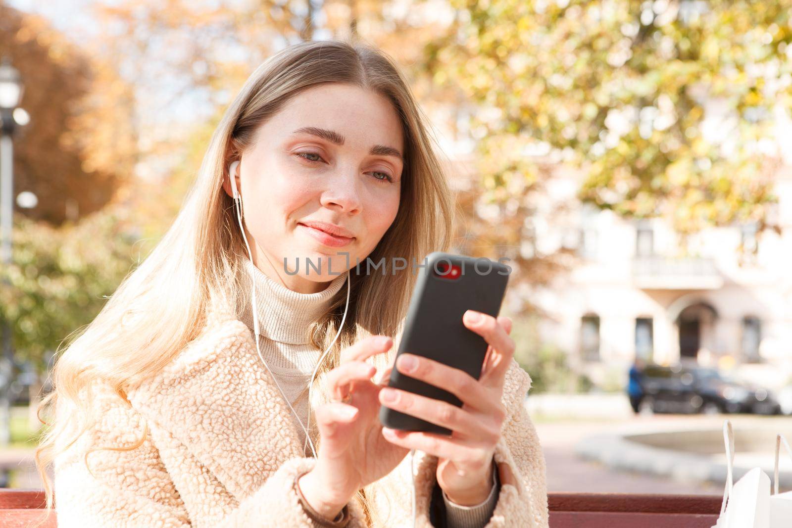 Beautiful woman smiling, using earphones with her smart phone in the park in autumn