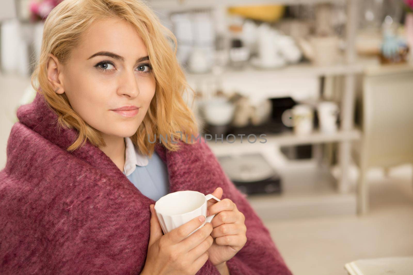 Attractive young woman smiling to the camera holding cup of coffee sitting comfortably wrapped in warm woolen blanket home recreation weekend lifestyle living leisure beauty serenity homeware