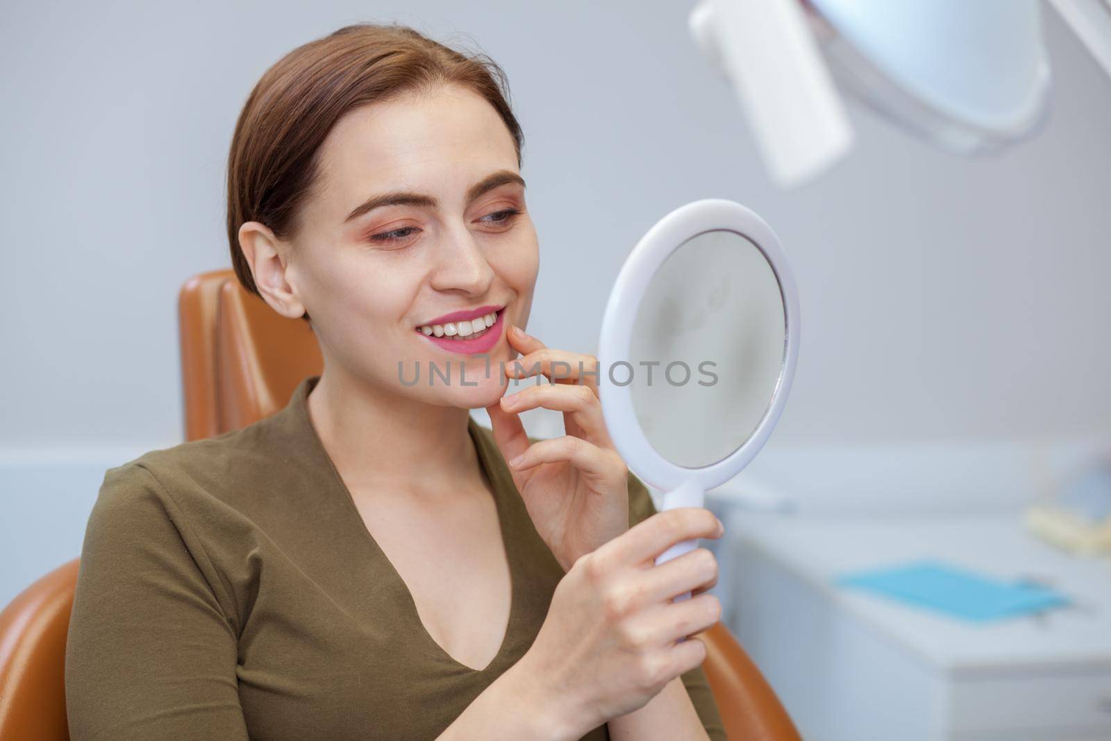 Attractive young woman smiling, examining her teeth in the mirror, while visiting dentist. Woman enjoying result of professional teeth cleaning and whitening, copy space. Health, wellness concept