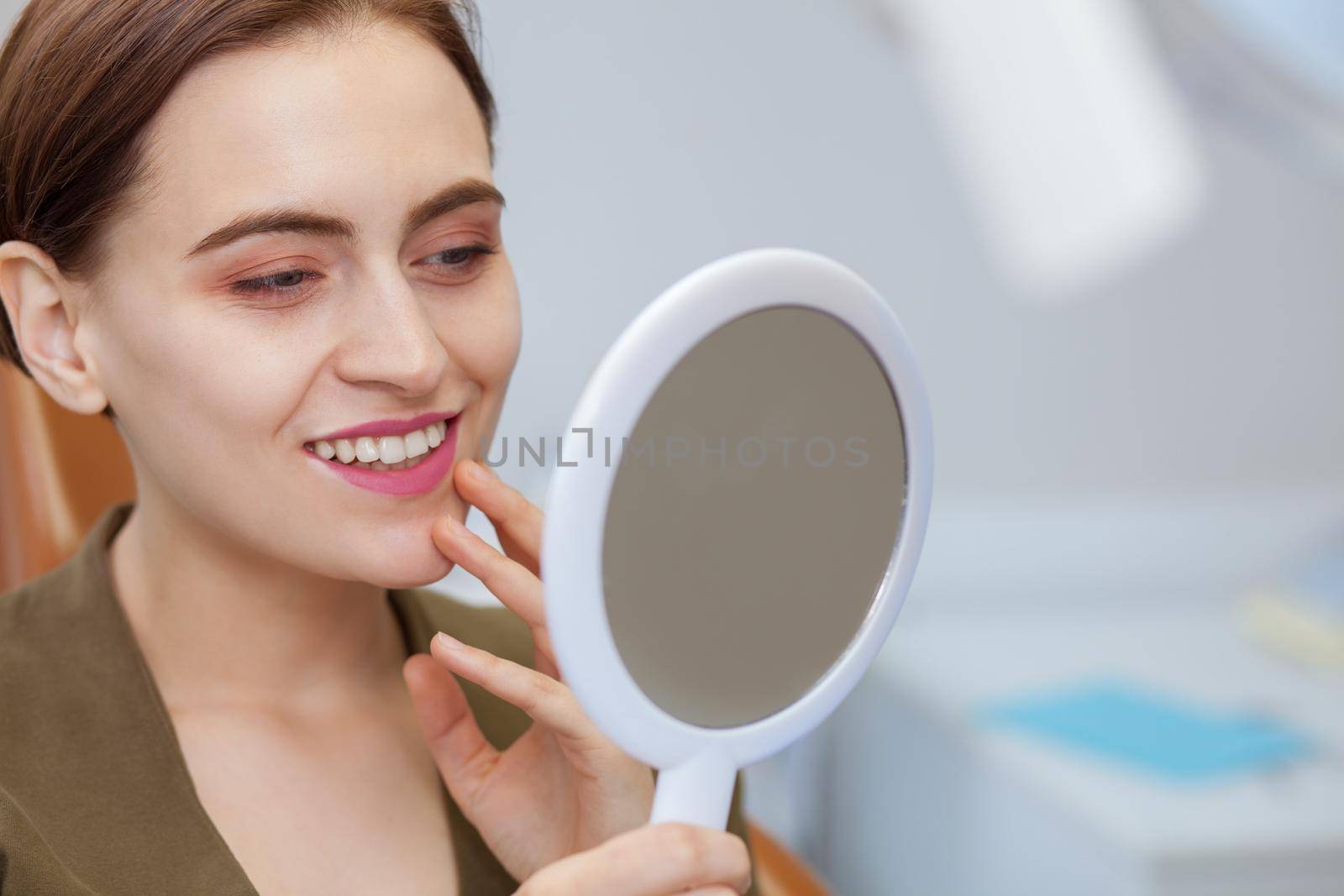Close up of a young beautiful woman smiling, looking in the mirror at the dental clinic. Attractive female patient examining her perfect whitened smile after medical treatment, Dental health concept